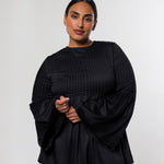 A woman modeling a black pleated long-sleeve top with dramatic bell sleeves and beige trousers, standing against a light gray studio background.