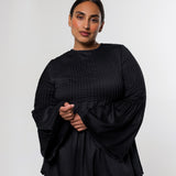 A woman modeling a black pleated long-sleeve top with dramatic bell sleeves and beige trousers, standing against a light gray studio background.
