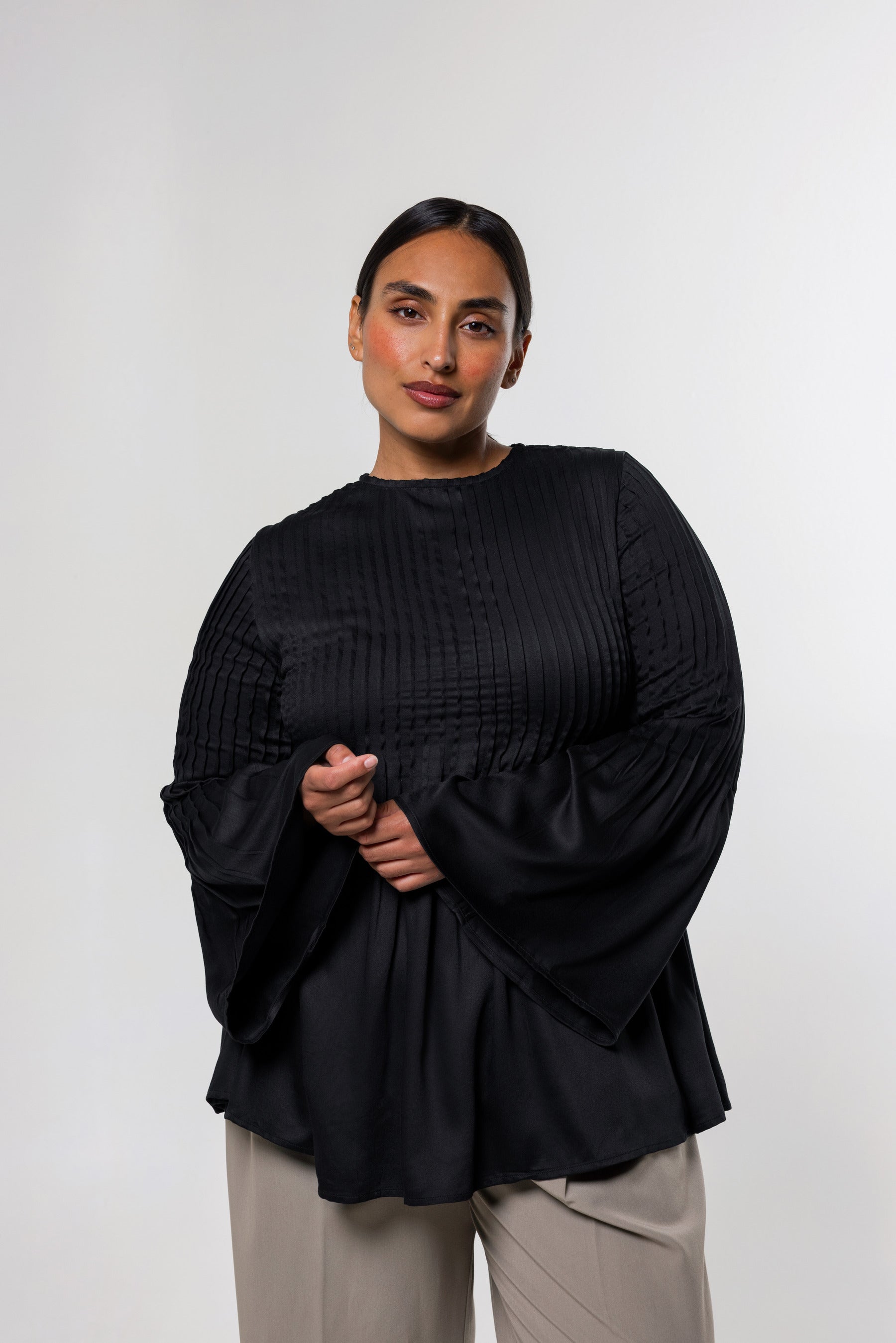 A woman modeling a black pleated long-sleeve top with dramatic bell sleeves and beige trousers, standing against a light gray studio background.