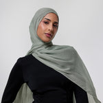 A woman wearing a light sage green hijab and a black one-shoulder dress, photographed against a neutral light gray studio background.