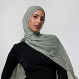 A woman wearing a light sage green hijab and a black one-shoulder dress, photographed against a neutral light gray studio background.
