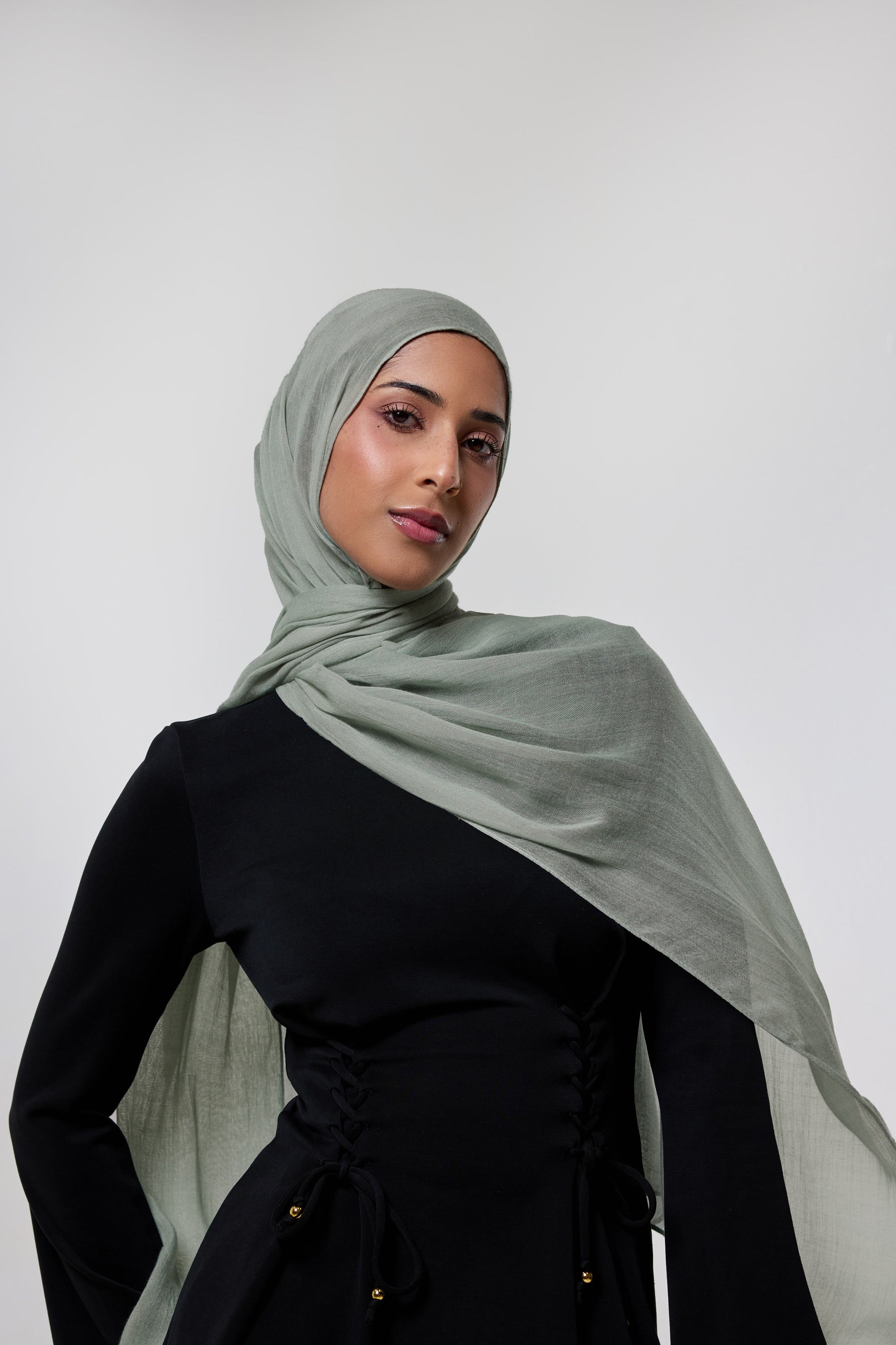 A woman wearing a light sage green hijab and a black one-shoulder dress, photographed against a neutral light gray studio background.