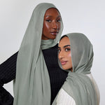 Two women wearing sage green hijabs, posed against a plain light gray studio background. One wears a full-length draped hijab over a black top, and the other wears a shorter side-drape hijab over a white top.