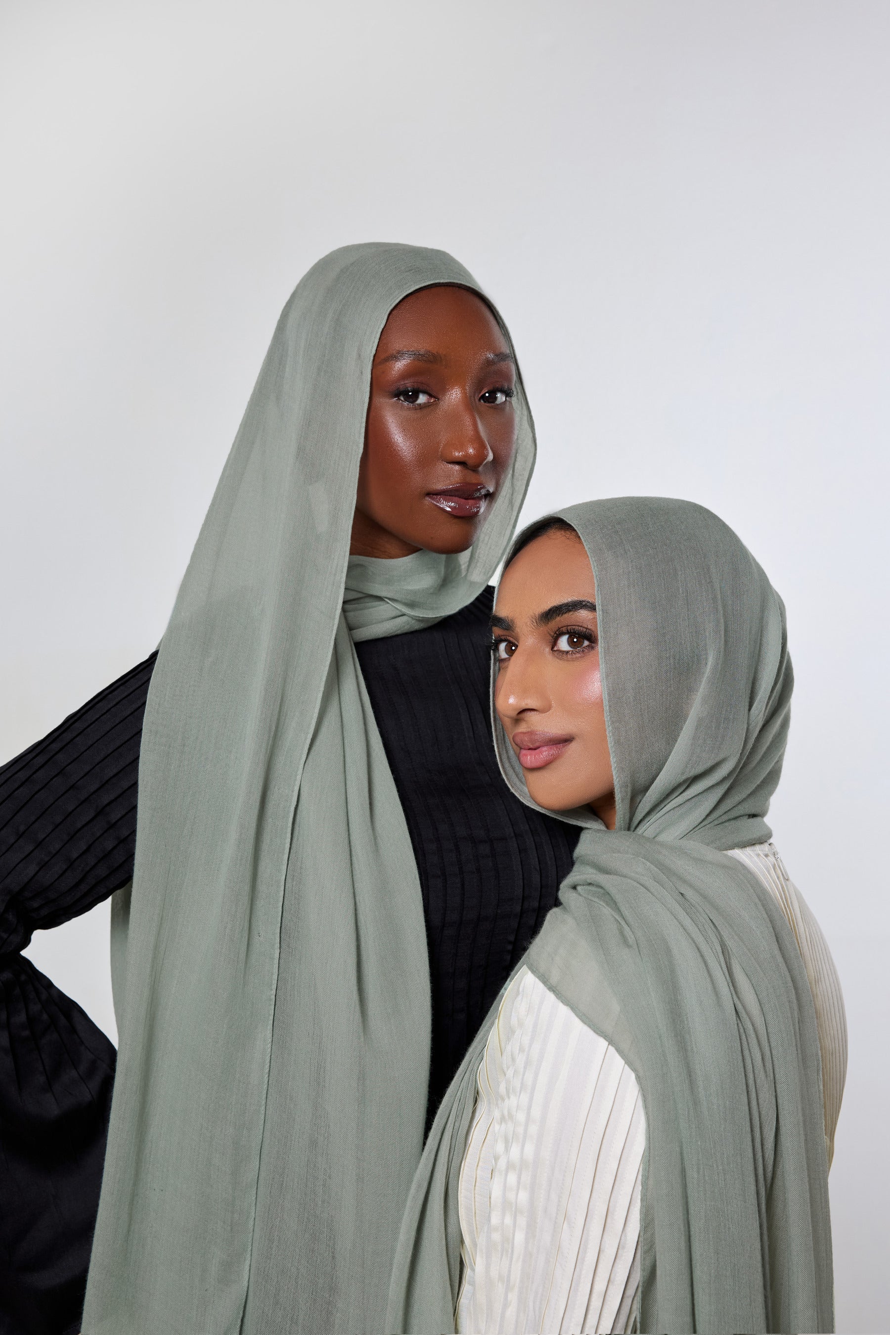 Two women wearing sage green hijabs, posed against a plain light gray studio background. One wears a full-length draped hijab over a black top, and the other wears a shorter side-drape hijab over a white top.