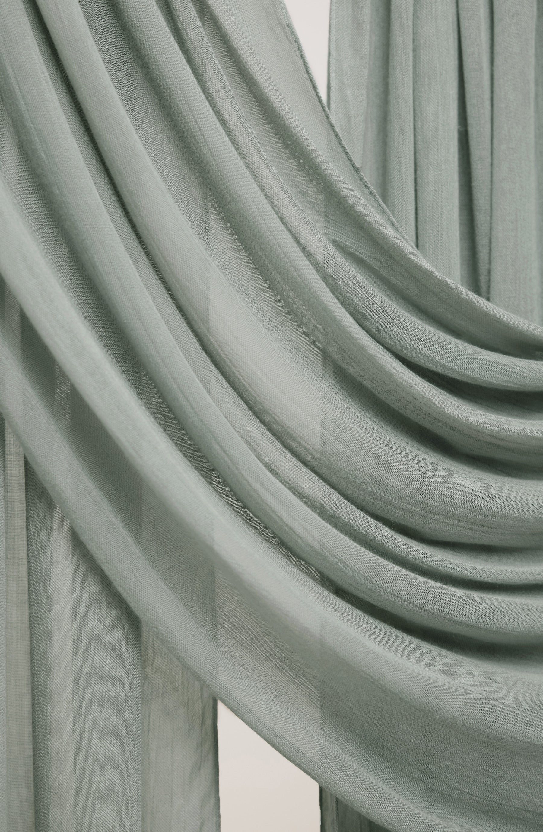 A close-up of draped sage green sheer curtains with soft folds and a light neutral background visible in the gaps.