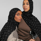 Two women wearing black hijabs with white patterns against a gray background
