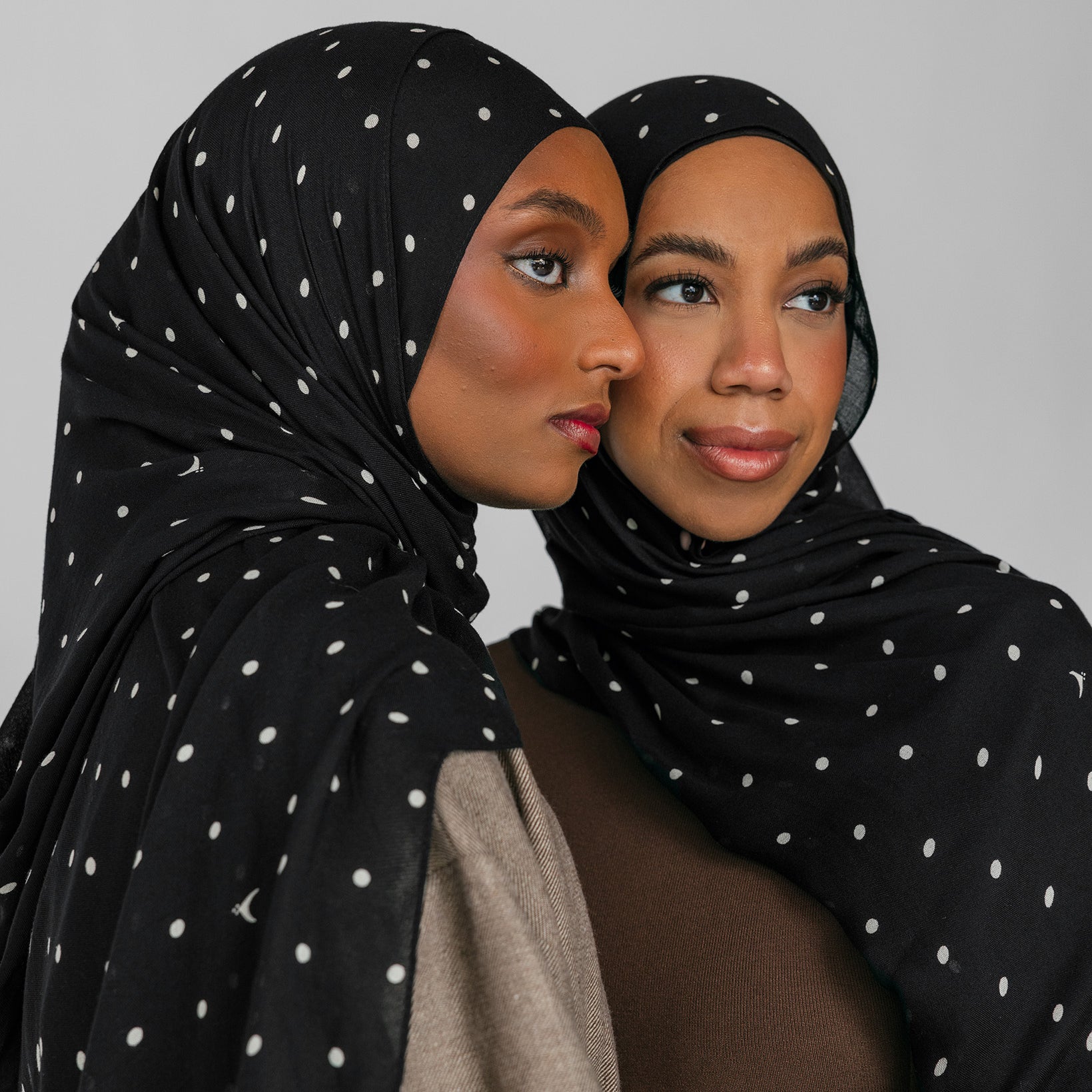 Two women wearing black polka-dot hijabs against a gray background