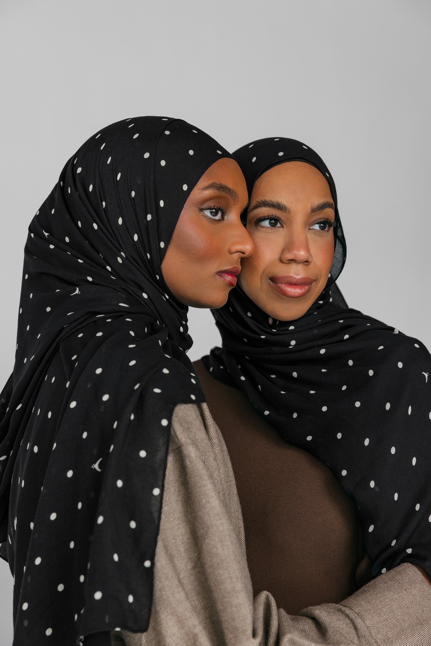 Two women wearing black polka-dot hijabs against a gray background