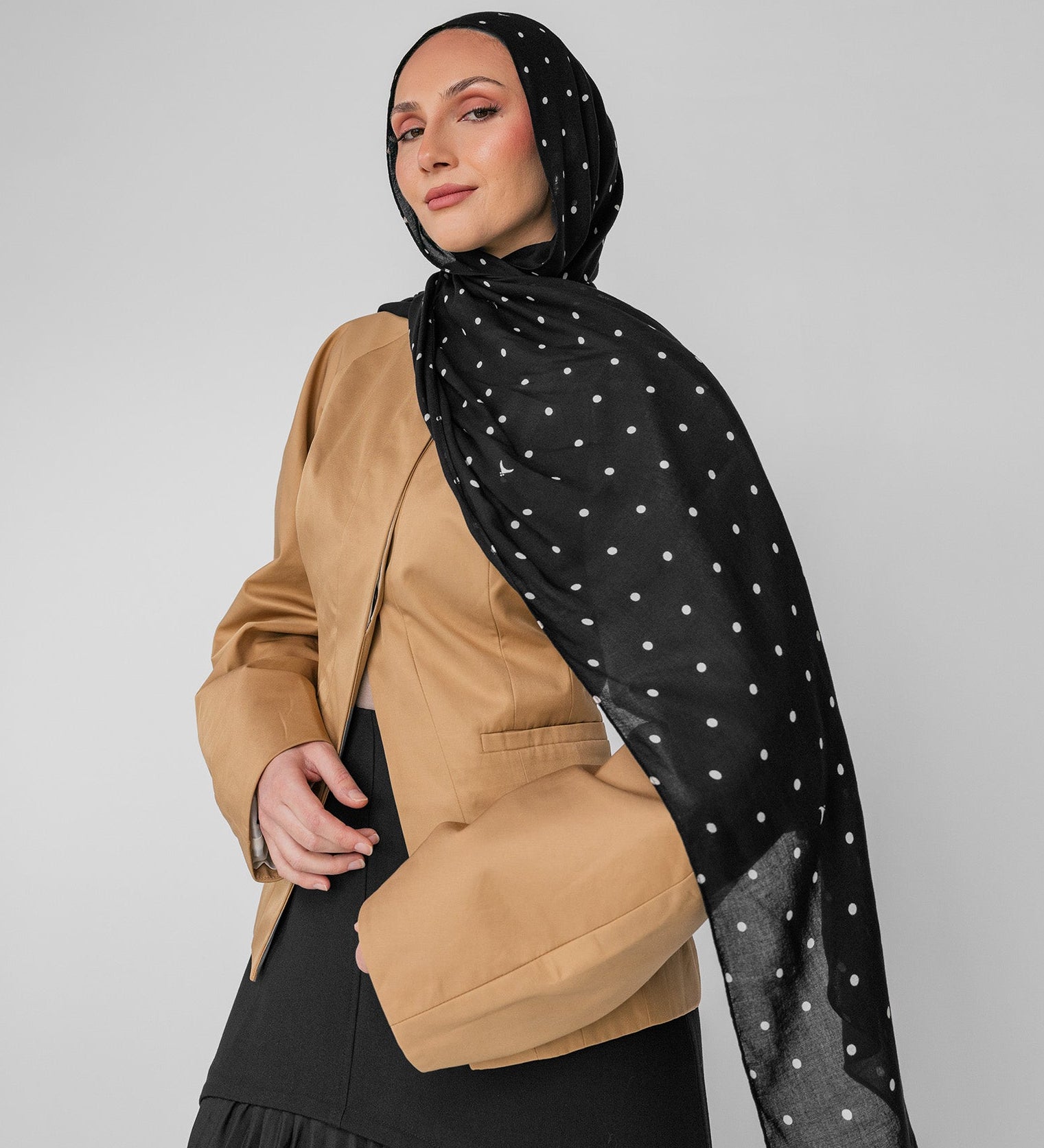 Woman wearing a black polka-dot hijab and a beige top on a gray background