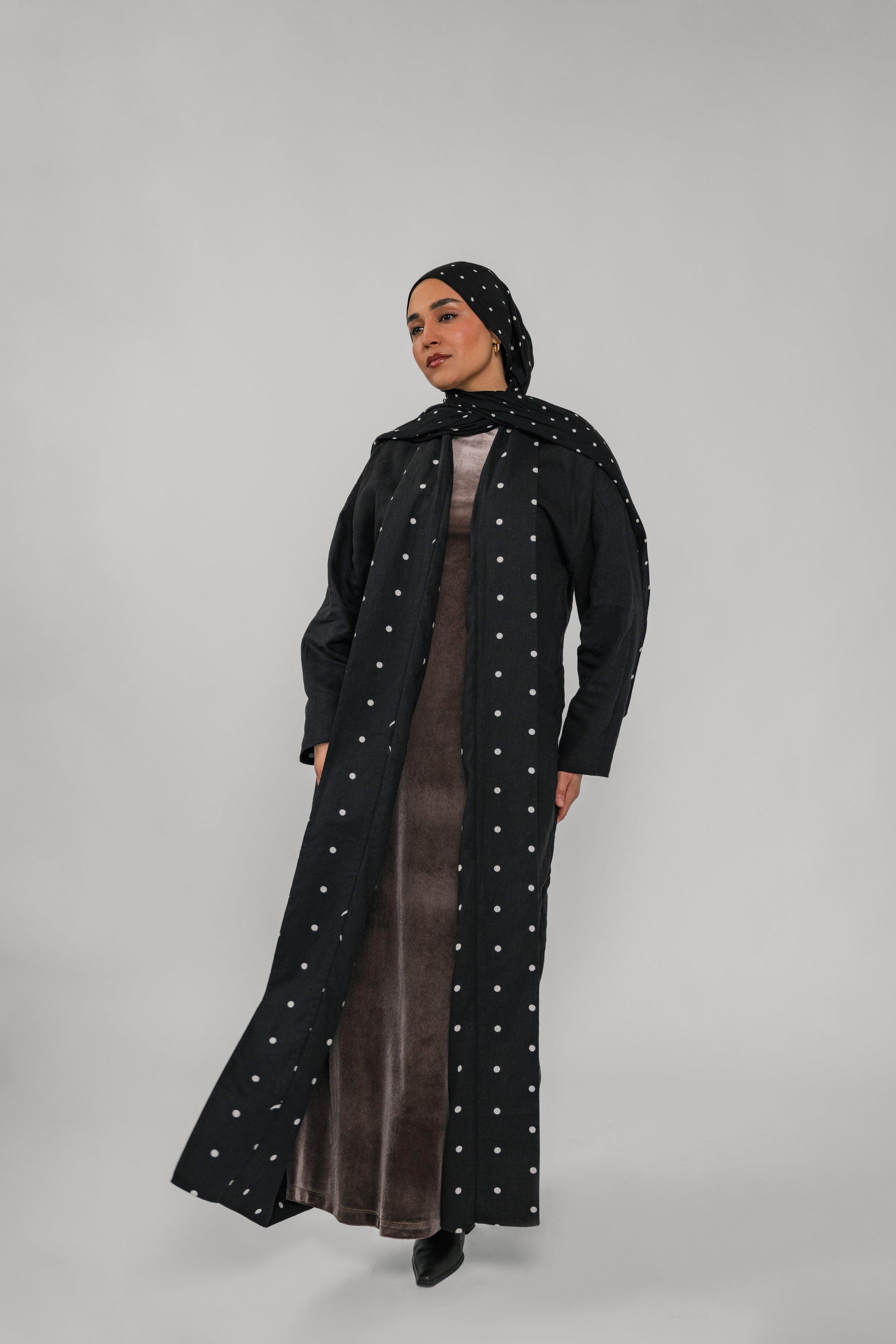 Woman wearing a long black coat with white polka dots and a matching headscarf on a gray background