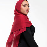 A woman wearing a vibrant red chiffon hijab and a black abaya, photographed from the side against a clean white studio background.