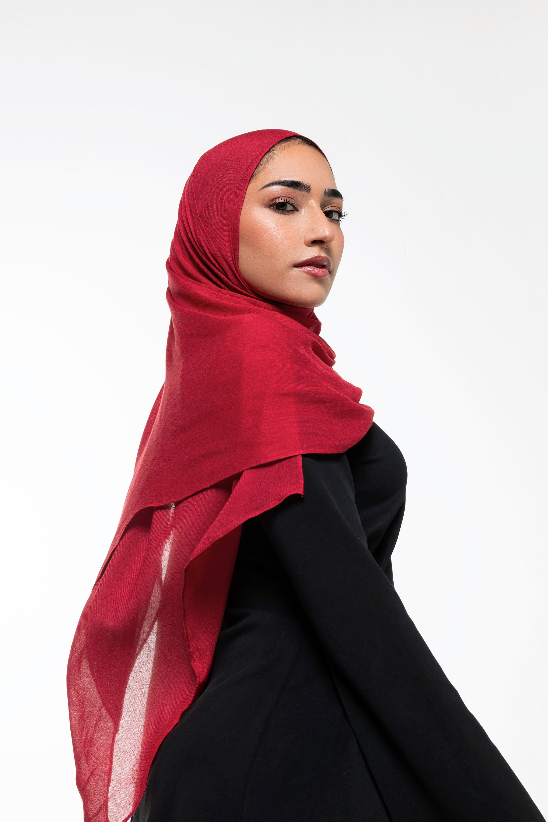 A woman wearing a vibrant red chiffon hijab and a black abaya, photographed from the side against a clean white studio background.