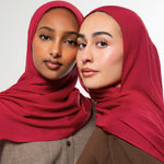 Two women wearing matching deep red hijabs, posed closely against a clean light gray studio background.
