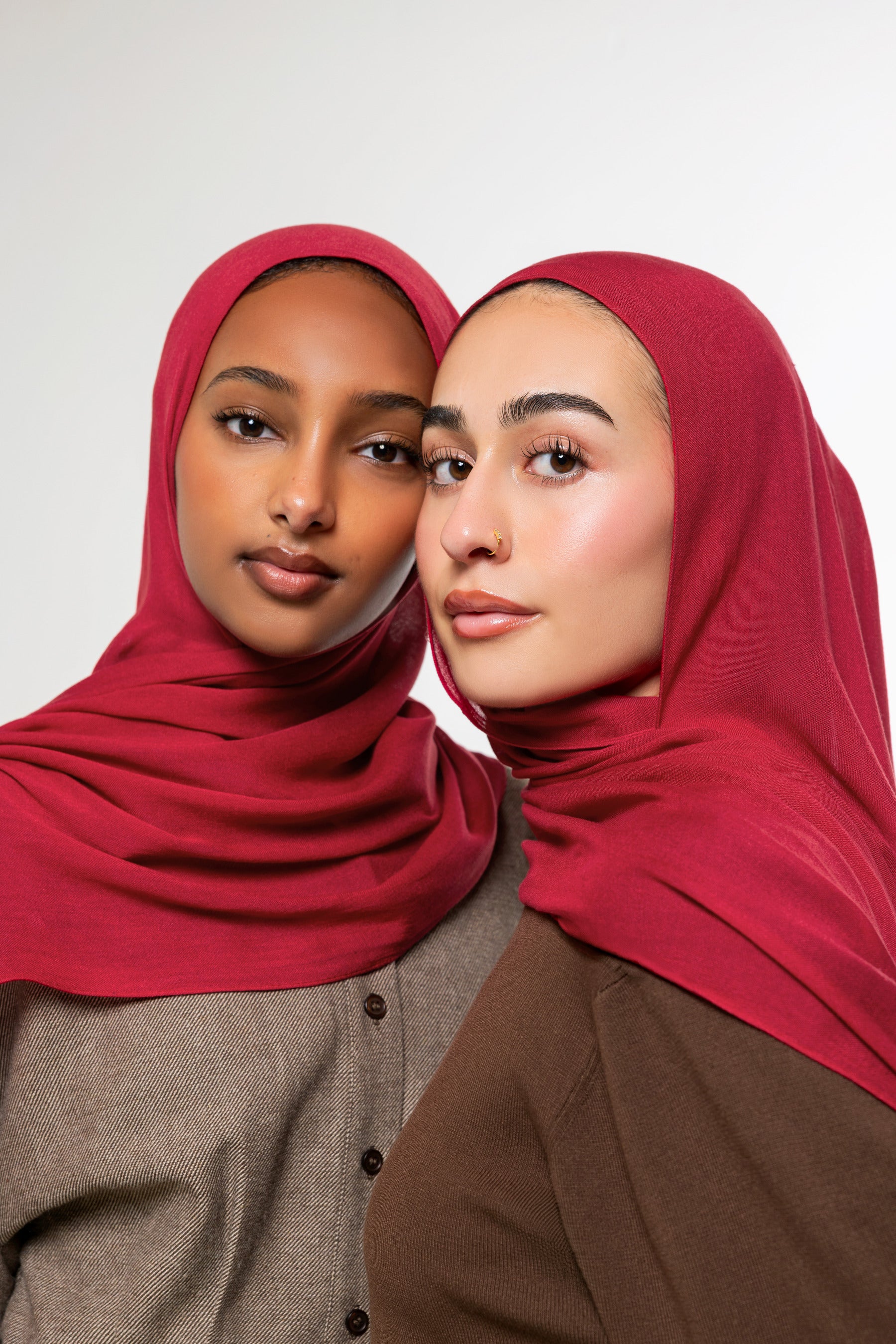 Two women wearing matching deep red hijabs, posed closely against a clean light gray studio background.