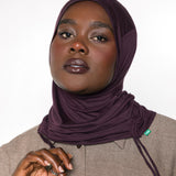 A woman wearing a deep plum purple hijab and a textured taupe-beige button-up shirt, photographed against a clean white studio background.