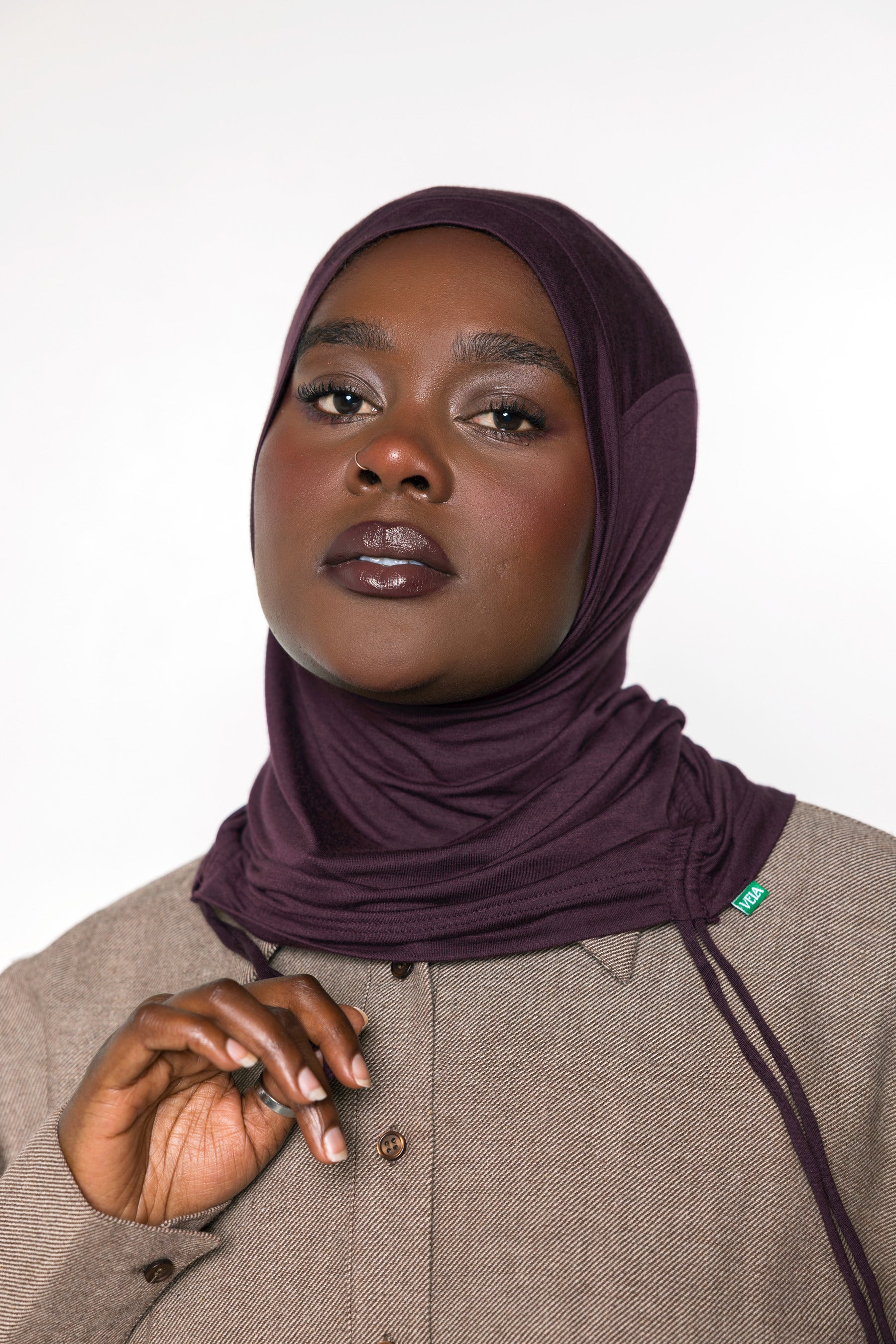 A woman wearing a deep plum purple hijab and a textured taupe-beige button-up shirt, photographed against a clean white studio background.