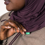 Close-up of a person wearing a deep plum purple hijab and a taupe-brown textured jacket, with a small green VEAIA brand tag visible near the collar.