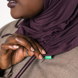 Close-up of a person wearing a deep plum purple hijab and a taupe-brown textured jacket, with a small green VEAIA brand tag visible near the collar.