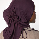 A woman shown in profile wearing a dark plum purple ruched hijab with drawstrings, paired with a textured beige top against a plain white background.