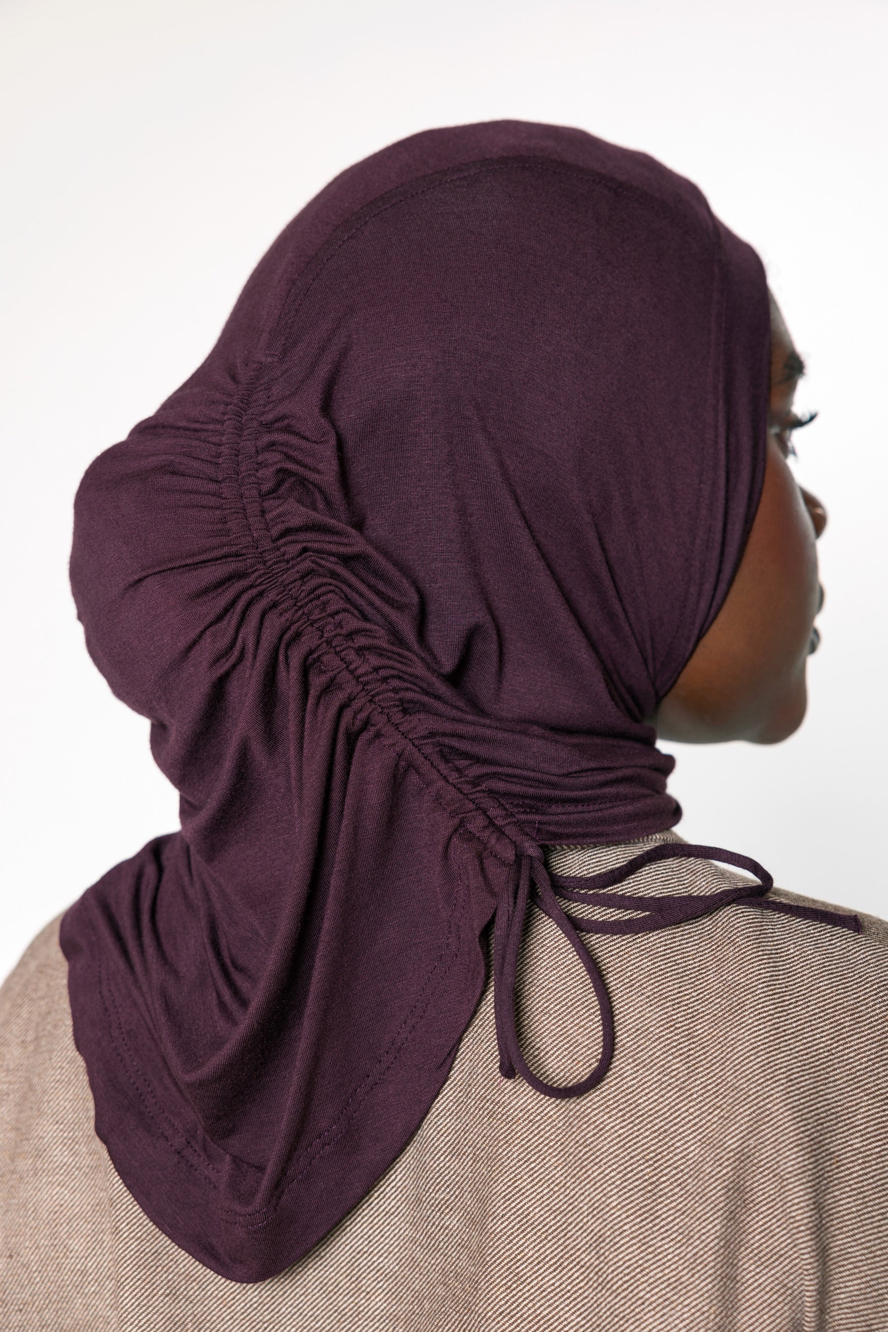 A woman shown in profile wearing a dark plum purple ruched hijab with drawstrings, paired with a textured beige top against a plain white background.