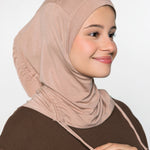 A woman wearing a soft, draped hijab in a dusty rose-beige color, paired with a dark brown top, shown against a plain light gray background.