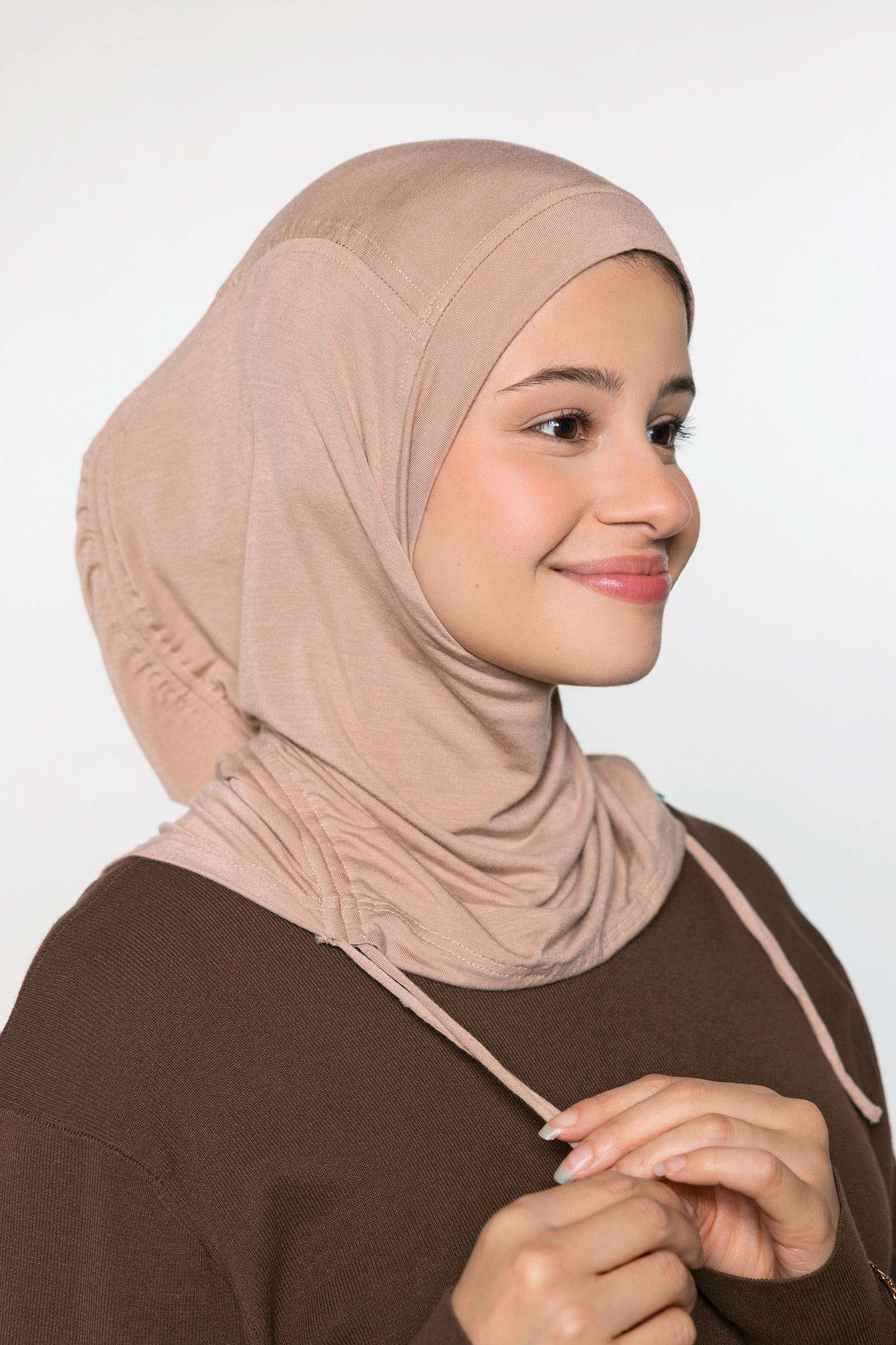 A woman wearing a soft, draped hijab in a dusty rose-beige color, paired with a dark brown top, shown against a plain light gray background.