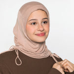 A woman wearing a light beige/tan hijab and a brown long-sleeve top, shown from the chest up against a plain light gray studio background.