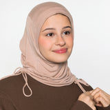 A woman wearing a light beige/tan hijab and a brown long-sleeve top, shown from the chest up against a plain light gray studio background.