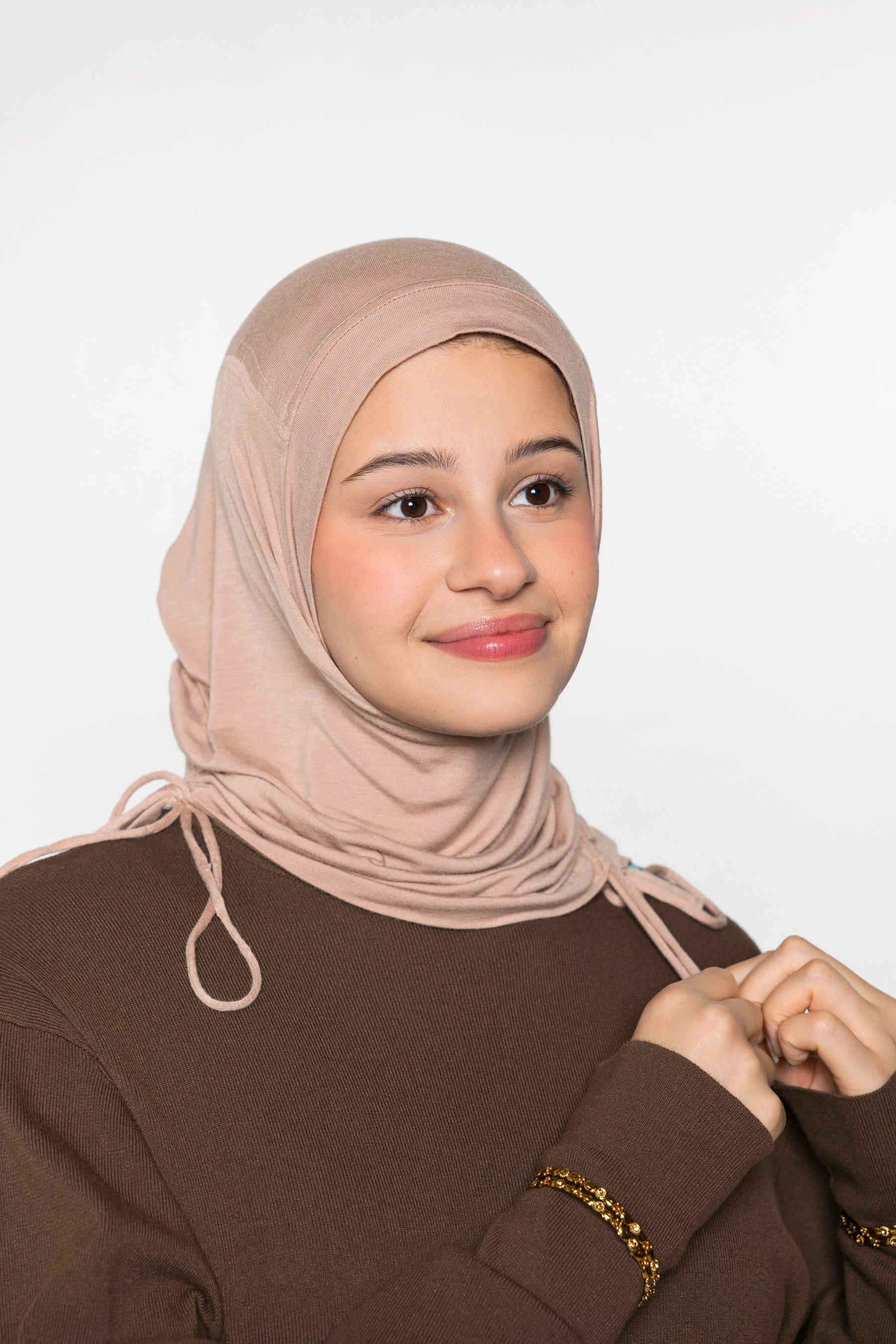 A woman wearing a light beige/tan hijab and a brown long-sleeve top, shown from the chest up against a plain light gray studio background.