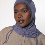 A woman wearing a slate blue-gray hijab and a textured beige button-up shirt, photographed against a neutral light gray background.