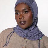 A woman wearing a slate blue-gray hijab and a textured beige button-up shirt, photographed against a neutral light gray background.