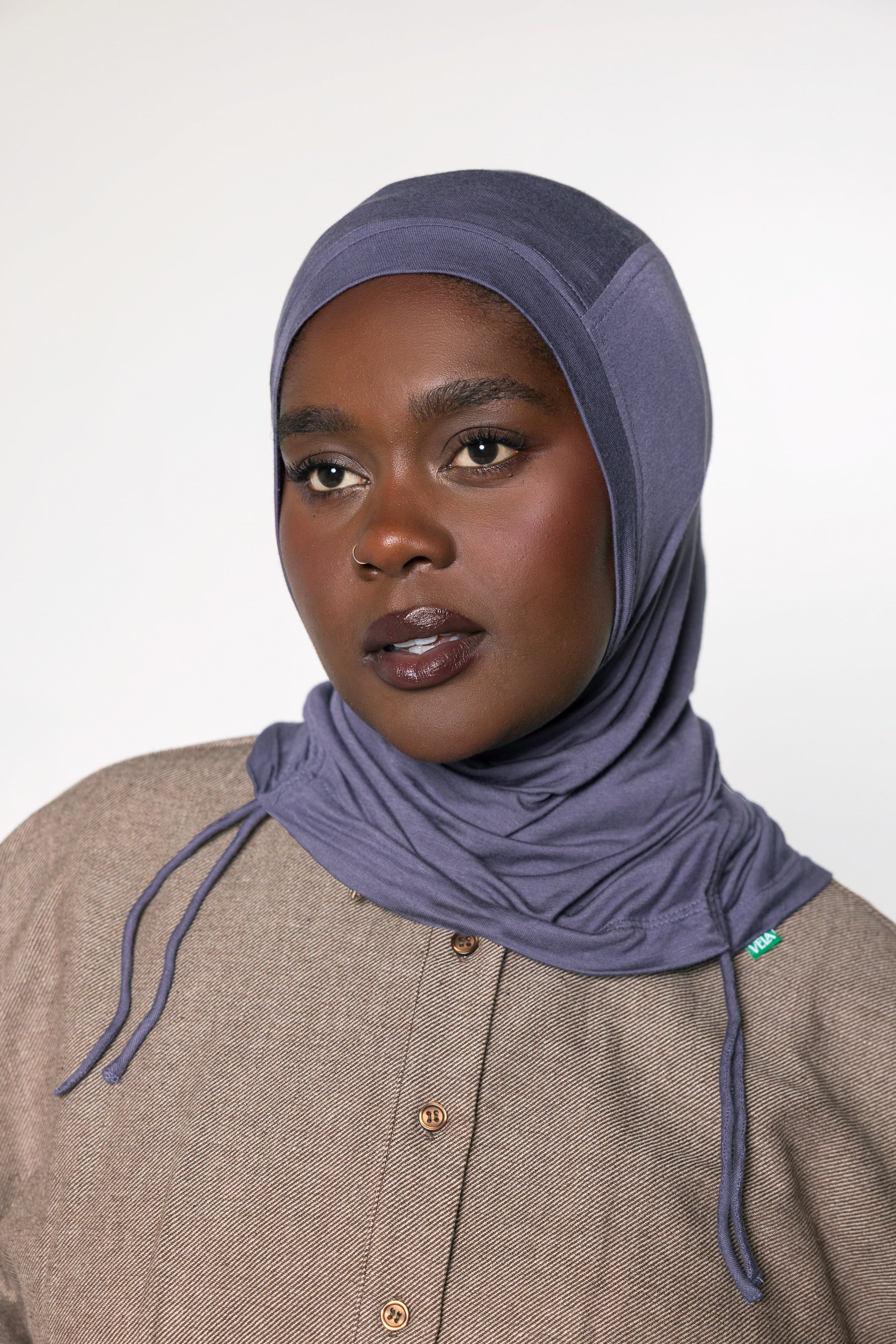 A woman wearing a slate blue-gray hijab and a textured beige button-up shirt, photographed against a neutral light gray background.