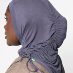 A side-profile view of a person wearing a dusty purple ruched hijab with drawstrings, paired with a brown textured jacket, against a plain light gray background.