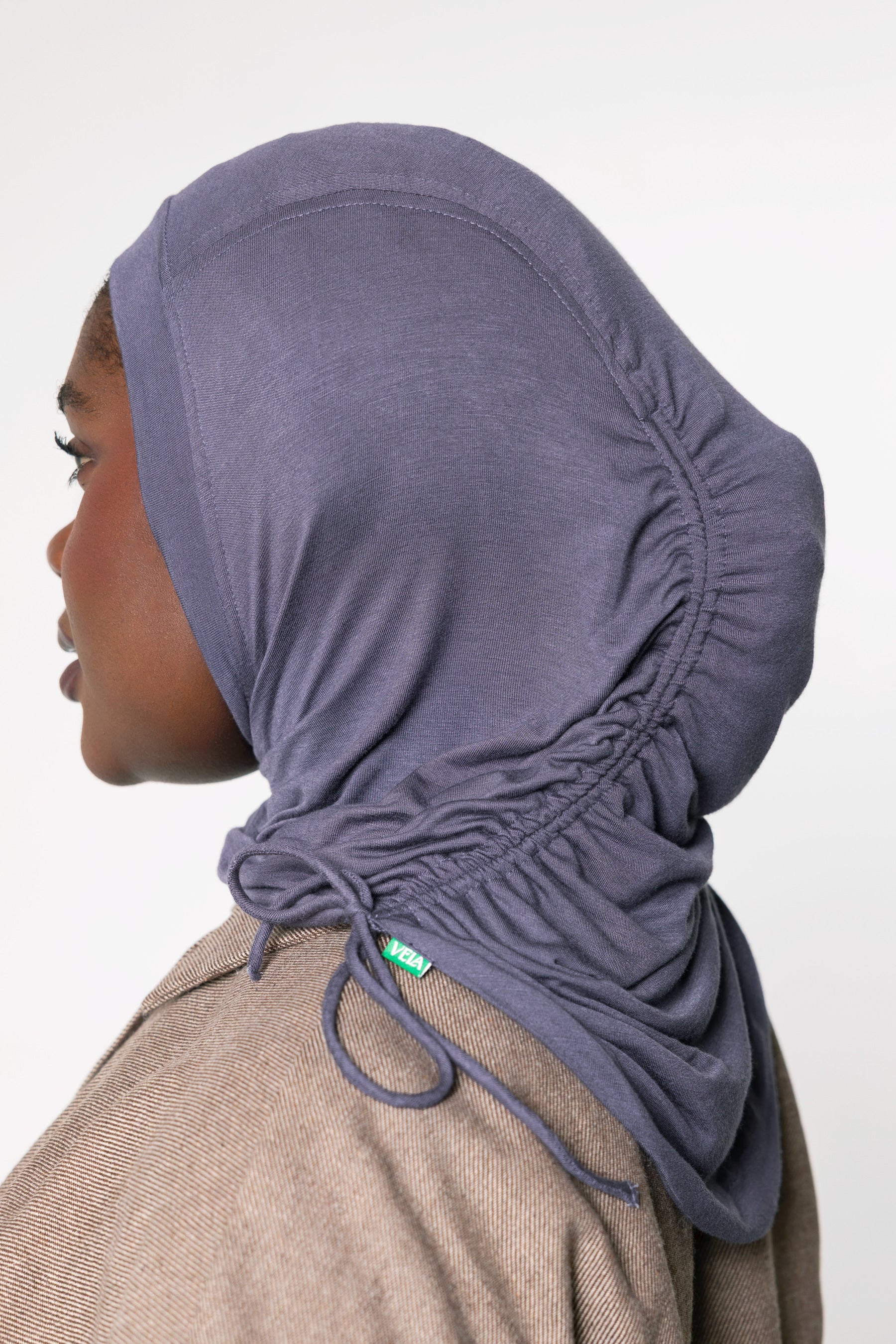 A side-profile view of a person wearing a dusty purple ruched hijab with drawstrings, paired with a brown textured jacket, against a plain light gray background.