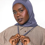 A woman wearing a slate blue-gray hijab and a taupe button-up shirt, adjusting the front of her hijab against a plain white background.