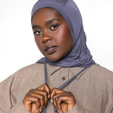 A woman wearing a slate blue-gray hijab and a taupe button-up shirt, adjusting the front of her hijab against a plain white background.