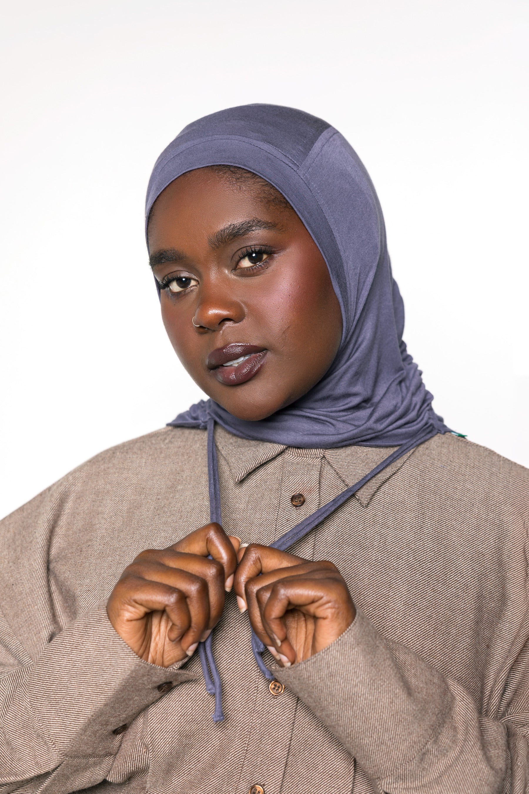 A woman wearing a slate blue-gray hijab and a taupe button-up shirt, adjusting the front of her hijab against a plain white background.
