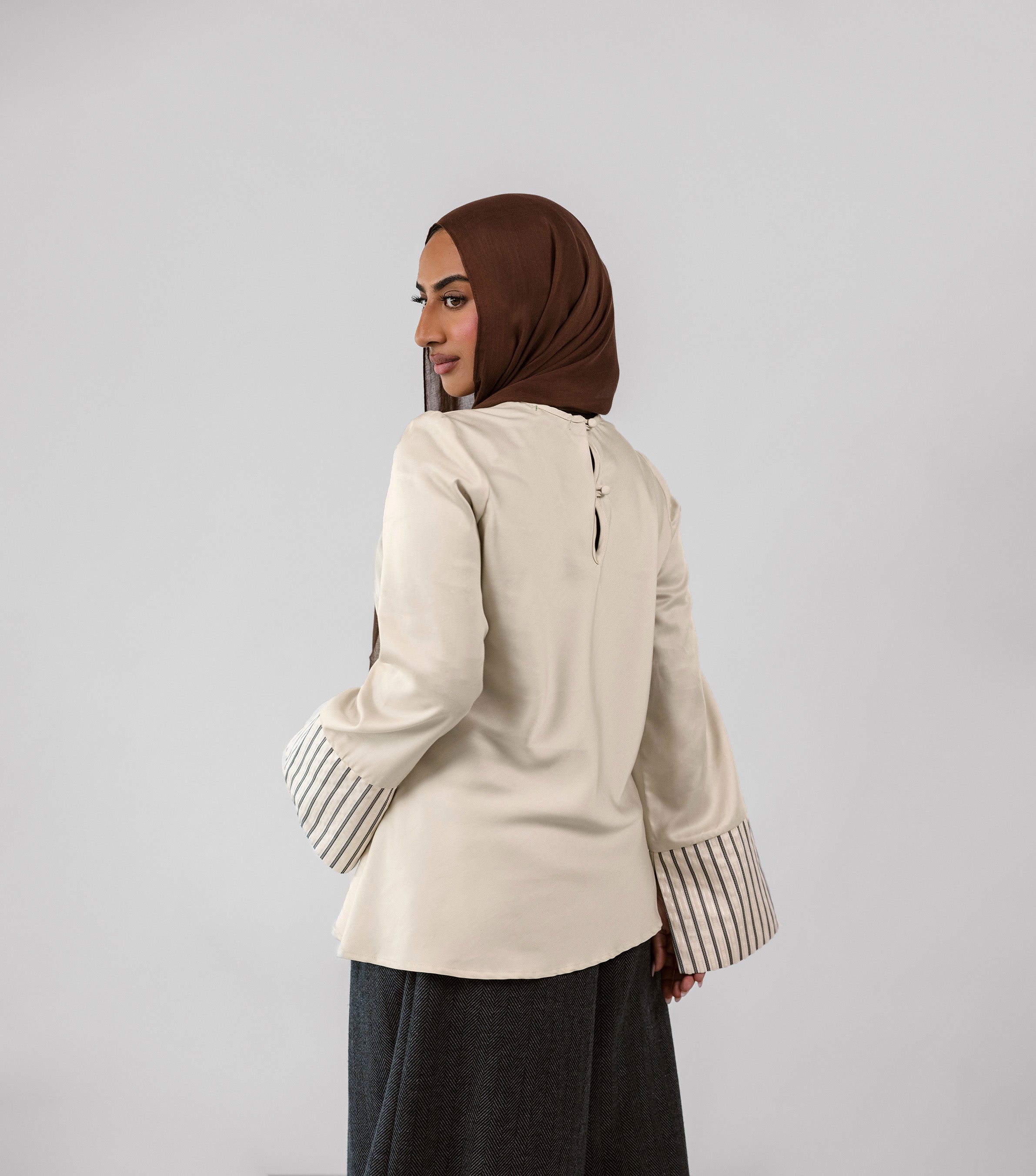 Woman wearing a beige jacket with striped cuffs and a brown hijab on a light gray background