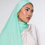 A woman wearing a light mint green hijab and a cream-colored draped top, photographed against a plain light gray studio background.
