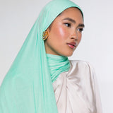 A woman wearing a light mint green hijab and a cream-colored draped top, photographed against a plain light gray studio background.