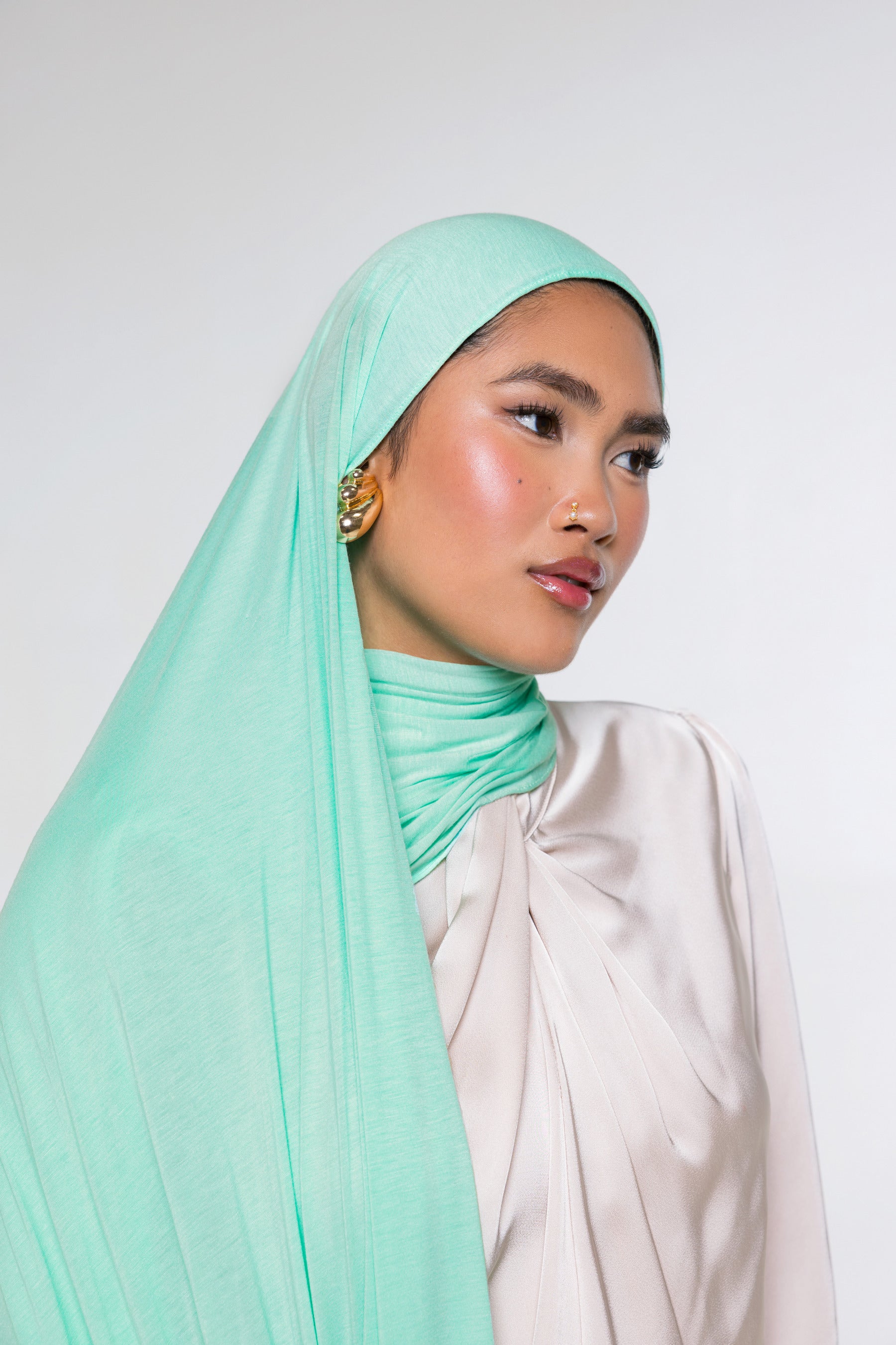 A woman wearing a light mint green hijab and a cream-colored draped top, photographed against a plain light gray studio background.
