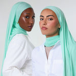 Two women wearing mint green hijabs and white button-up shirts, posed against a light gray studio background.