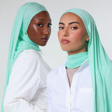 Two women wearing mint green hijabs and white button-up shirts, posed against a light gray studio background.