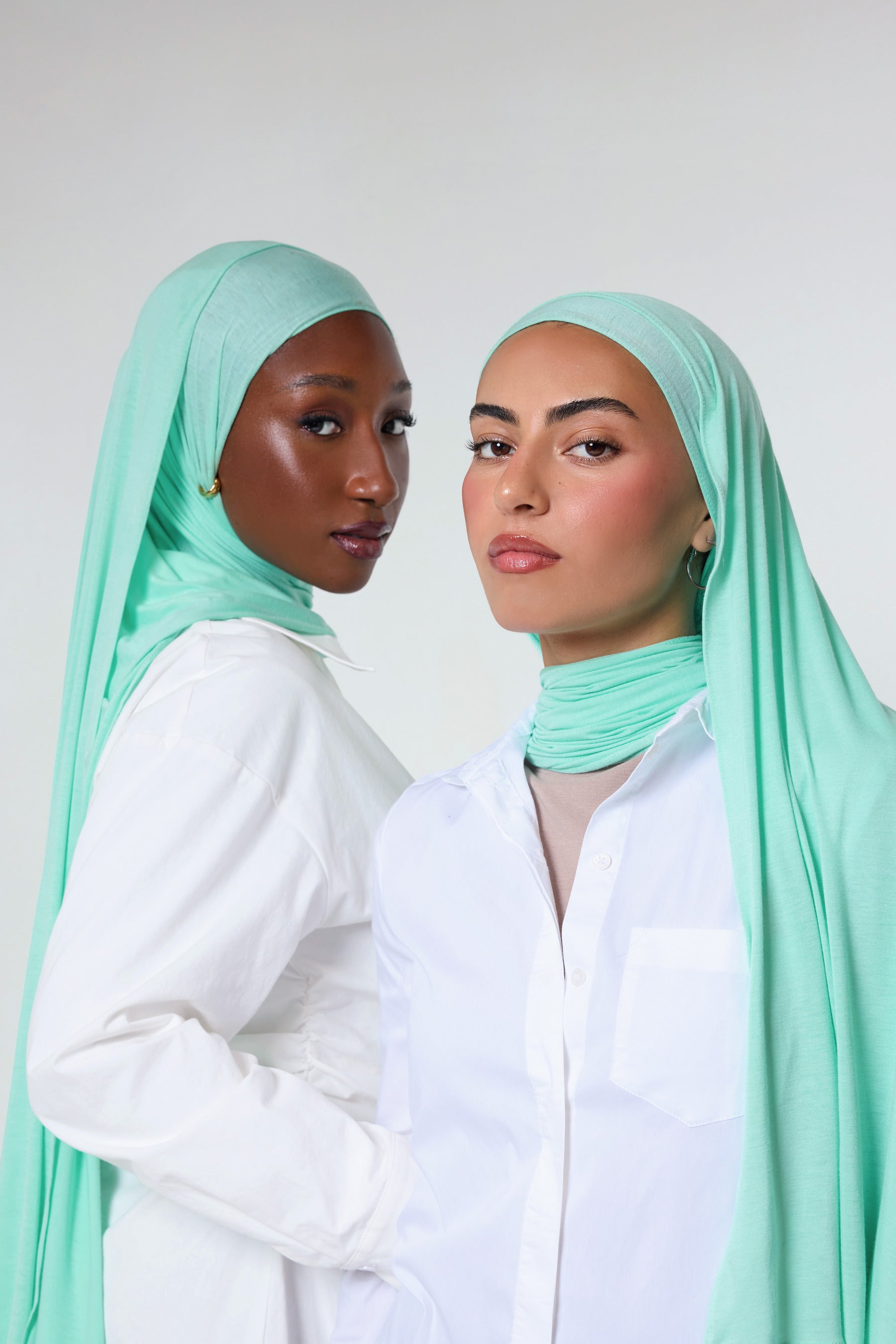 Two women wearing mint green hijabs and white button-up shirts, posed against a light gray studio background.