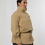 A woman wearing a beige high-neck, long-sleeve jacket with gathered detailing and a drawstring waist, paired with dark gray pants, photographed against a plain light gray studio background.