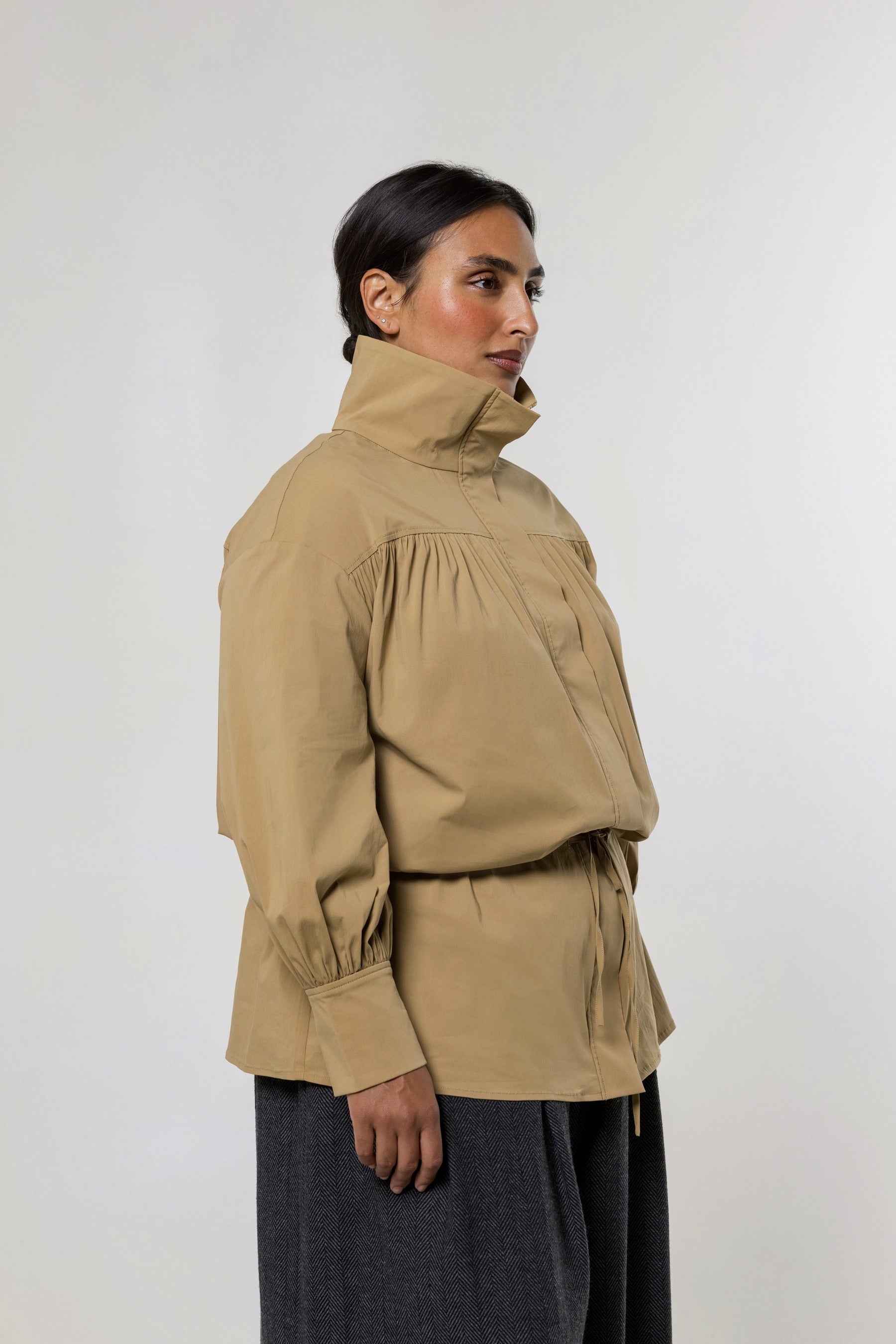 A woman wearing a beige high-neck, long-sleeve jacket with gathered detailing and a drawstring waist, paired with dark gray pants, photographed against a plain light gray studio background.