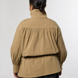 A model viewed from the back wearing a tan high-neck long-sleeve jacket with gathered detailing and dark gray wide-leg trousers, set against a plain light gray studio background.