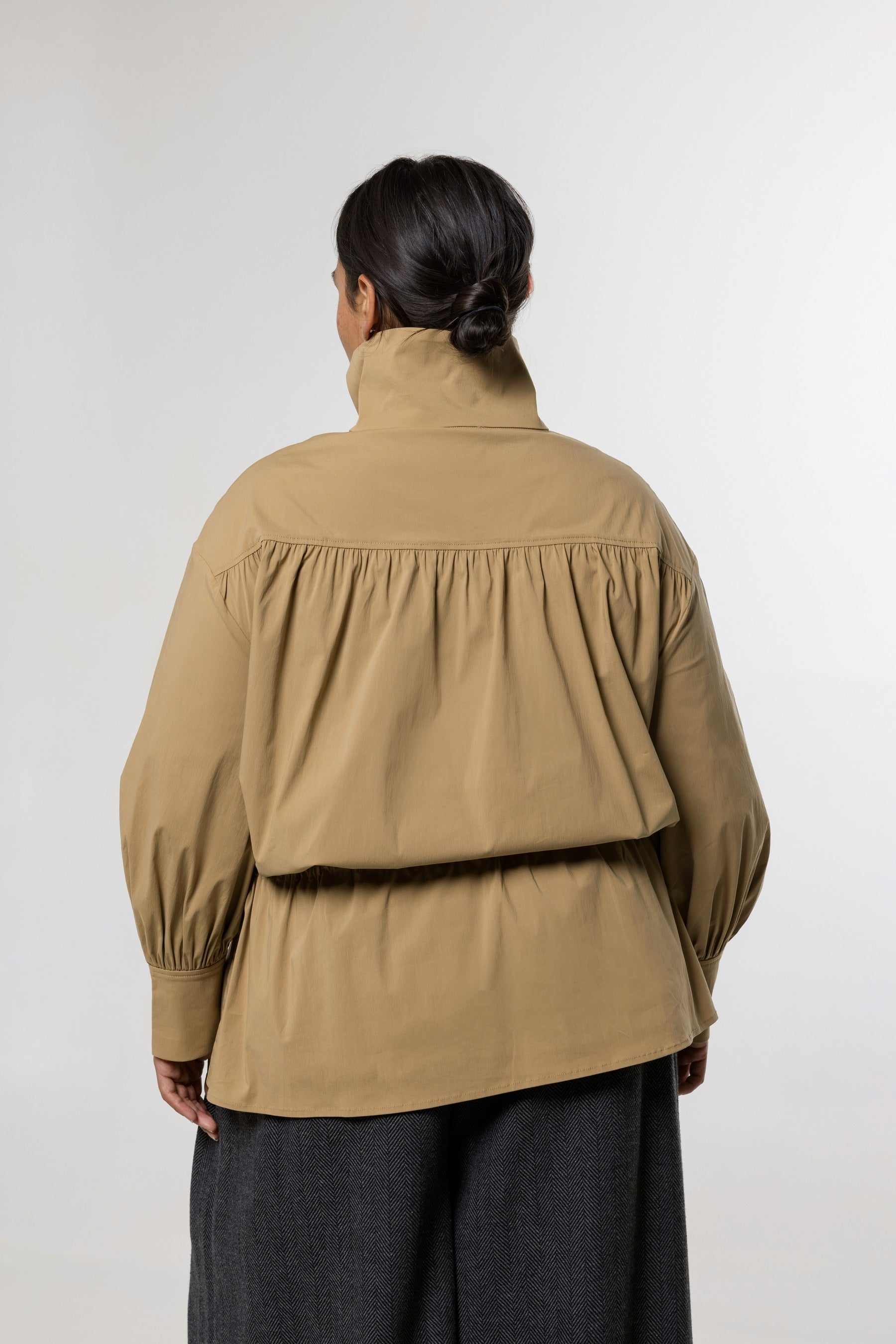 A model viewed from the back wearing a tan high-neck long-sleeve jacket with gathered detailing and dark gray wide-leg trousers, set against a plain light gray studio background.