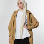 A woman modeling a tan hooded jacket over a white top and black pants, wearing a light beige hijab, against a plain light gray studio background.