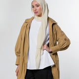 A woman modeling a tan hooded jacket over a white top and black pants, wearing a light beige hijab, against a plain light gray studio background.